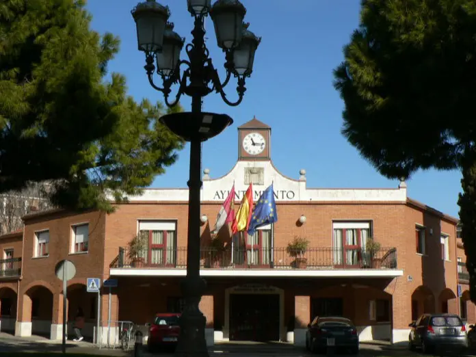 Edificio de ladrillo del ayuntamiento de Azuqueca de Henares, con un reloj en la fachada. Hay banderas de España, Castilla-La Mancha y la Unión Europea. Árboles y farolas rodean el edificio. 