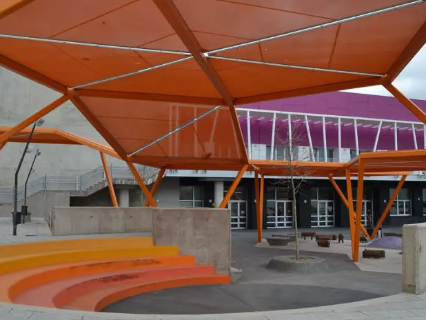 La imagen muestra una estructura moderna en el Centro Joven de Azuqueca de Henares. Tiene un techo naranja geométrico sostenido por pilares del mismo color. Hay gradas circulares en tonos de amarillo a rojo y un edificio con detalles púrpuras al fondo.