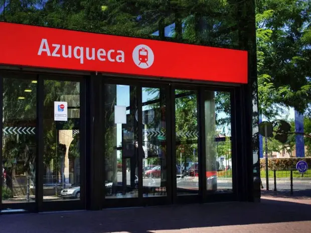 La imagen muestra la entrada de la estación de Renfe de Azuqueca de Henares. Un cartel rojo con el texto "Azuqueca" y un ícono de tren está sobre las puertas de vidrio. Al fondo, hay árboles y señales de tráfico