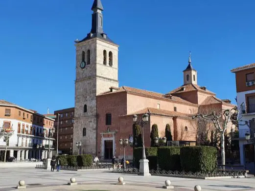 La imagen muestra una plaza amplia con una iglesia de ladrillo en el centro, destacando su torre con reloj y campanario. Alrededor hay edificios residenciales y personas caminando. El cielo está despejado. 