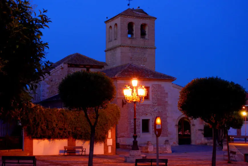 La imagen muestra la iglesia de San Miguel de Azuqueca de Henares al anochecer. La iglesia tiene una torre cuadrada de ladrillo con campanario. Está iluminada por farolas, rodeada de árboles y un banco en primer plano